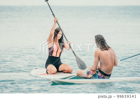 Sea woman and man on sup. Silhouette of happy young woman and man, surfing on SUP board, confident paddling through water surface. Idyllic sunset. Active lifestyle at sea or river. 99639141
