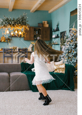 A girl with long hair in a white dress is spinning in the living room, with a Christmas tree and kitchen in the background A girl with long hair in a white dress is spinning in the living room, with a Christmas tree and kitchen in the background 99645834