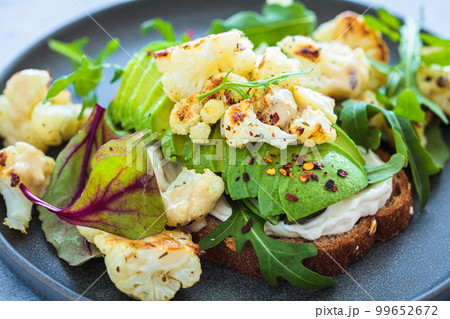 Vegan toast with avocado, baked cauliflower, lettuce and tahini on dark plate. Healthy plant based organic diet. 99652672
