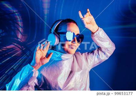 Portrait of a smiling dancing woman in sunglasses and headphones in neon light. Music lover. Silent disco. Woman in holographic clothes on the background of music vibes background. Futuristic Party. Portrait of a smiling dancing woman in sunglasses and headphones in neon light. Music lover. Silent disco. Woman in holographic clothes on the background of music vibes background. Futuristic Party. 99653177