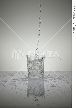 Vertical shot of water pouring into crystal glass with droplets around, on a white shiny table Vertical shot of water pouring into crystal glass with droplets around, on a white shiny table 99657770