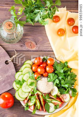 Fresh slices of cucumbers, tomatoes, sesame seeds in a bowl and parsley leaves on a plate on a wooden table. Oil and seasoning for cooking vegetable salad. Top view Fresh slices of cucumbers, tomatoes, sesame seeds in a bowl and parsley leaves on a plate on a wooden table. Oil and seasoning for cooking vegetable salad. Top view 99659485