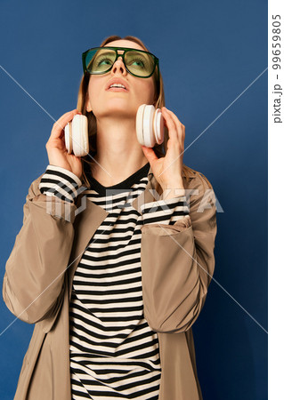 Adorable caucasian young woman in wireless headphones listening to music and dancing on blue navy background. Blonde girl in coat smiles. 99659805