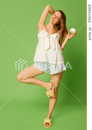 Beautiful cheerful woman wearing comfortable pyjamas, homewear eats sweet cake and rejoices isolated on green background. 99659868