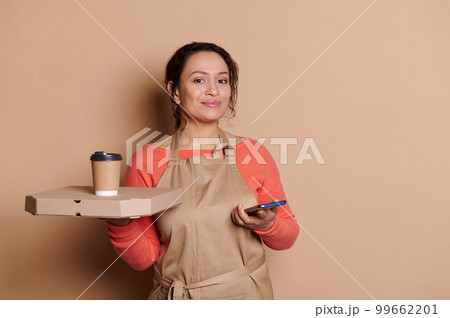 Charming Latin American positive female bartender, waitress in chef's apron, posing with takeaway fast food and mobile phone, isolated on beige backdrop. Online food ordering. Delivery service concept 99662201