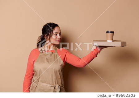 Attractive African American woman waitress, barkeeper, wearing beige apron, holding a cardboard flat box with tasty Italian pizza and takeaway hot drink in eco paper cup, isolated on cream background Attractive African American woman waitress, barkeeper, wearing beige apron, holding a cardboard flat box with tasty Italian pizza and takeaway hot drink in eco paper cup, isolated on cream background 99662203