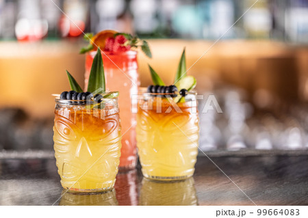 Mai tai cocktails in hawaiian glasses garnished with pineapple, blueberries and strawberries on bar counter. Mai tai cocktails in hawaiian glasses garnished with pineapple, blueberries and strawberries on bar counter. 99664083