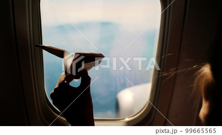 close-up. Silhouette of a child's hand with small paper plane against the background of airplane window. Child sitting by aircraft window and playing with little paper plane. during flight on airplane 99665565
