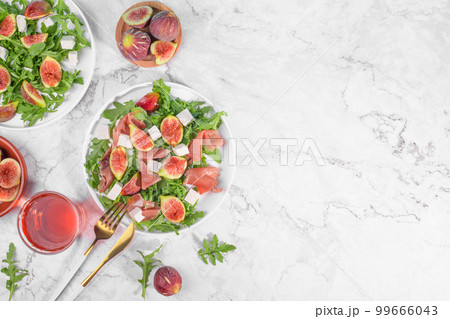 Autumn figs salad with arugula, feta, raw ham in two white plate on light marlbe background served with rose wine. 99666043