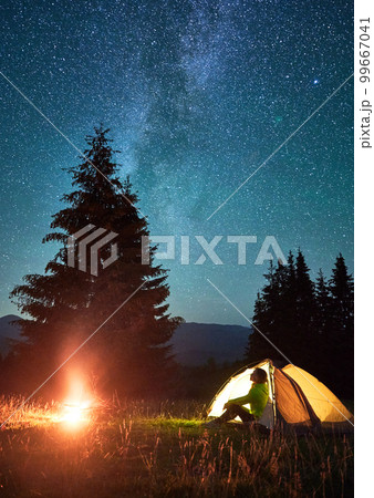 Night camping in mountains under starry sky and Milky way. Female tourist sitting in tent entrance in campsite, admiring landscape and burning campfire. Concept of traveling and hiking. 99667041