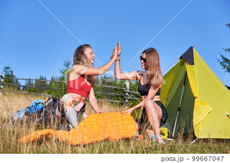 Two women hikers camping in mountains. Young female tourists giving high five in summer, smiling, enjoying. Concept of tourism and hiking. 99667047