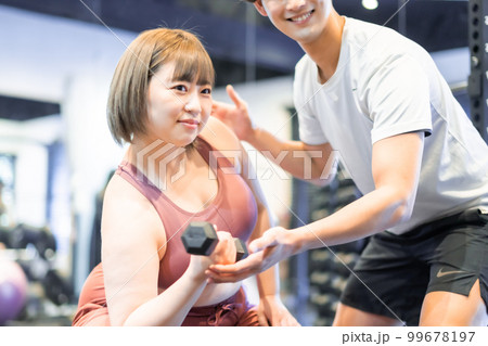 スポーツジムでトレーナーに指導を受ける笑顔の若い女性 99678197