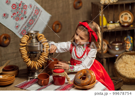 Little girl in folk dress pours tea from samovar while sitting in folk decor room. Shrovedite Little girl in folk dress pours tea from samovar while sitting in folk decor room. Shrovedite 99678754