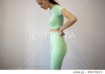 Slender young woman in a light turquoise tracksuit with leggings and top looks down and pulls up her pants 99679923