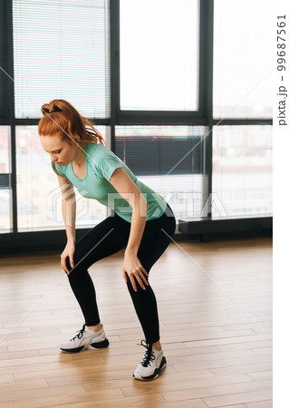Vertical shot of athletic young woman doing squats exercising and stop training as feeling sharp knee pain during fitness workout at home by window. Female athlete sportswoman touching leg in injury. 99687561