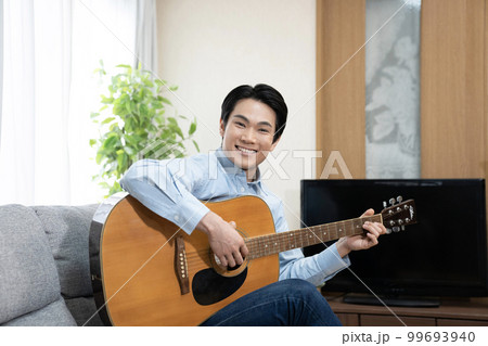 ソファーに座ってギターを弾いて微笑む男性 99693940