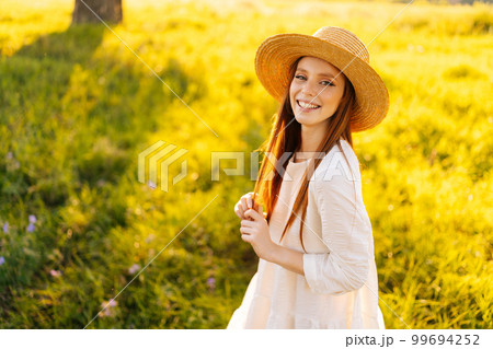 Portrait of lovely redhead young woman in straw hat and white dress standing posing on beautiful meadow of green grass looking at camera, on background of warm sunlight at summer day. 99694252