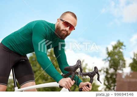 A professional cyclist in sportswear with glasses is preparing for a morning bike ride in a green city park. The concept of an active and healthy lifestyle. A professional cyclist in sportswear with glasses is preparing for a morning bike ride in a green city park. The concept of an active and healthy lifestyle. 99694546