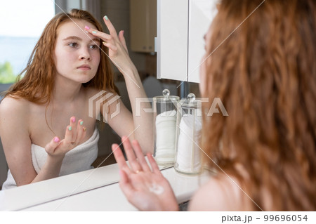 Red-haired girl applies cream to her face looking at herself in the bathroom mirror after shower Red-haired girl applies cream to her face looking at herself in the bathroom mirror after shower 99696054