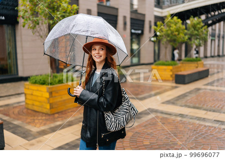 Medium shot of positive redhead young woman in fashion hat walking on European city street with transparent umbrella enjoying rainy weather outdoors, smiling looking away. Concept of female lifestyle. Medium shot of positive redhead young woman in fashion hat walking on European city street with transparent umbrella enjoying rainy weather outdoors, smiling looking away. Concept of female lifestyle. 99696077