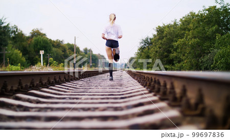 Beautiful sexy athletic young blond woman in top and shorts, running, jogging, Rear view. On the railway, on rails, sleepers, in the summer. Beautiful sexy athletic young blond woman in top and shorts, running, jogging, Rear view. On the railway, on rails, sleepers, in the summer. 99697836