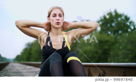 Beautiful sexy athletic young blond woman in black leggings performing exercises to make strong press, abdominal muscles. Sits on sleepers, on railway, in summer, Slowmotion 99697840
