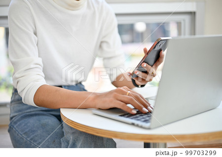 an Asian woman using her smartphone and laptop, remote working at the coffee shop. an Asian woman using her smartphone and laptop, remote working at the coffee shop. 99703299