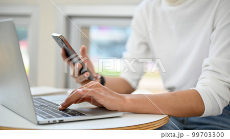 Cropped image of a female remote working at the cafe, using laptop and her phone. Cropped image of a female remote working at the cafe, using laptop and her phone. 99703300