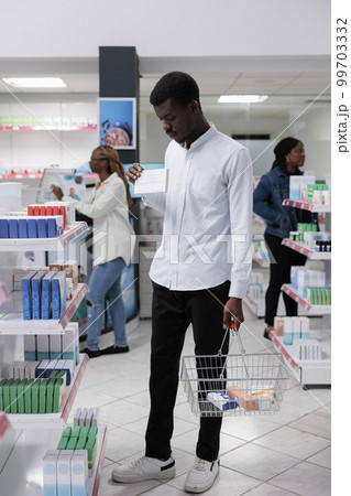 African american man shopping in drugstore, buying medications, reading instruction on tablets package, standing in pharmacy aisle. Client choosing vitamins, purchasing medicaments, wide shot African american man shopping in drugstore, buying medications, reading instruction on tablets package, standing in pharmacy aisle. Client choosing vitamins, purchasing medicaments, wide shot 99703332