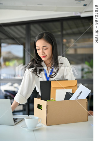Frustrated Asian female office employee packing her belongings at her desk, quitting job. 99703829