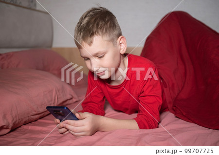 A cute boy in red pajamas is lying in bed under a blanket and playing on the phone A cute boy in red pajamas is lying in bed under a blanket and playing on the phone 99707725