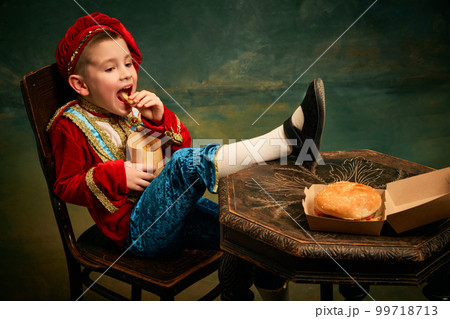 Happy cheerful little boy wearing costume of medieval page boy and prince eating hamburger with french fries over dark green background. Happy cheerful little boy wearing costume of medieval page boy and prince eating hamburger with french fries over dark green background. 99718713
