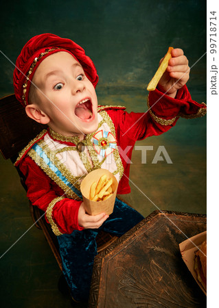 Happy cheerful little boy wearing costume of medieval page boy and prince eating hamburger with french fries over dark green background. Happy cheerful little boy wearing costume of medieval page boy and prince eating hamburger with french fries over dark green background. 99718714