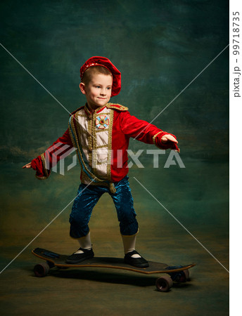 Smiling little boy dressed up as medieval little prince and pageboy skateboarding over dark vintage style background. Action, leisure activities, emotions, sport concept Smiling little boy dressed up as medieval little prince and pageboy skateboarding over dark vintage style background. Action, leisure activities, emotions, sport concept 99718735