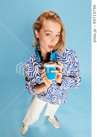 Tender cute young girl in headphones and casual clothes drinking coffee, posing over blue studio background. Delightful look Tender cute young girl in headphones and casual clothes drinking coffee, posing over blue studio background. Delightful look 99719796