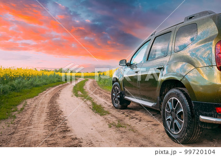 Car Renault Duster Or Dacia Duster Suv In Summer Rapeseed Field Countryside Landscape On Background Bright Orange Dramatic Sky. Summertime Flowering Canola, Rapeseed, Oilseed Field Meadow Grass 99720104