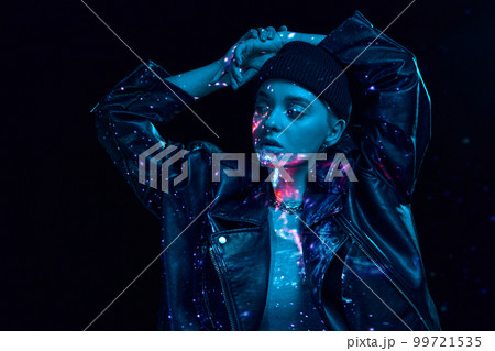 Portrait of young blonde girl with neon space holographic projection on body posing over dark background in blue neon lights. Cyberpunk style Portrait of young blonde girl with neon space holographic projection on body posing over dark background in blue neon lights. Cyberpunk style 99721535