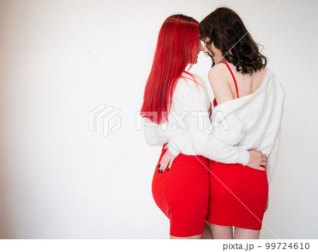 Rear view of two women dressed in identical red dresses and white sweaters. Lesbian intimacy. White background. 99724610