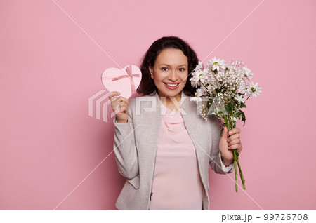 Cheerful middle-aged woman wearing elegant clothes, looking at camera with insightful gaze, showing bouquet of flowers and heart shaped gift box with happy surprise for women's Day, pink background Cheerful middle-aged woman wearing elegant clothes, looking at camera with insightful gaze, showing bouquet of flowers and heart shaped gift box with happy surprise for women's Day, pink background 99726708