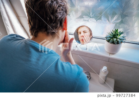 Concentrated caucasian teenage boy with acne problem take care his face skin at home. He looking in mirror and cleaning face with washing lotion in bathroom. Teenager skin care process 99727384