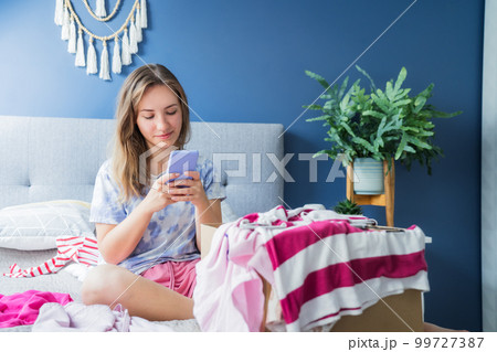 Smiling woman using smartphone to find places where to donate or sale clothes. Decluttering, Sorting clothes and Cleaning Up. Conscious consumer, sustainability. Reuse, second hand, Charity concept Smiling woman using smartphone to find places where to donate or sale clothes. Decluttering, Sorting clothes and Cleaning Up. Conscious consumer, sustainability. Reuse, second hand, Charity concept 99727387