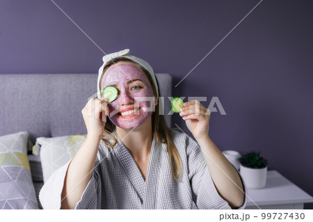 A young smiling woman with pink clay facial mask holds cucumber slices making a refreshing eye mask in bedroom. Natural cosmetic procedures for skin care at home. Beauty self-care. Selective focus. A young smiling woman with pink clay facial mask holds cucumber slices making a refreshing eye mask in bedroom. Natural cosmetic procedures for skin care at home. Beauty self-care. Selective focus. 99727430