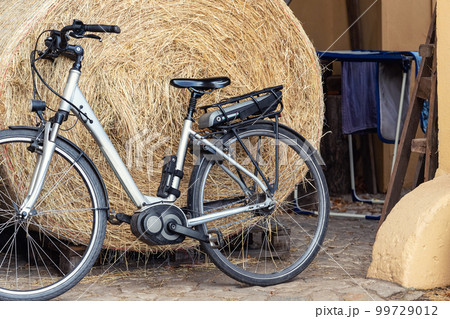 Electric eco green energy power hybrid e-bike parked against big hay straw bale at rural farm yard. Sustainable healthy countryside transport. Ebike at country village home Electric eco green energy power hybrid e-bike parked against big hay straw bale at rural farm yard. Sustainable healthy countryside transport. Ebike at country village home 99729012