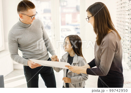 Father and daughter choosing glasses in optics store. Female optometrist helping customers choosing glasses. Little girl in optics store choosing glasses with her father. 99742950