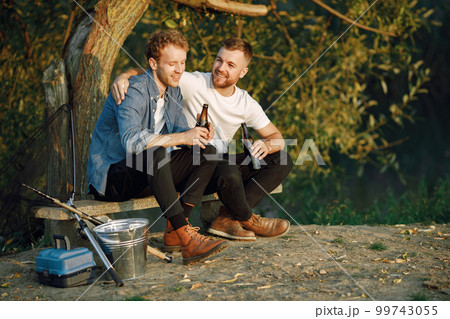 Friends is sitting near a tree and fishing. Two fishermen talking and drinking a beer. One man wearing white t-shirt and other blue shirt. Friends is sitting near a tree and fishing. Two fishermen talking and drinking a beer. One man wearing white t-shirt and other blue shirt. 99743055