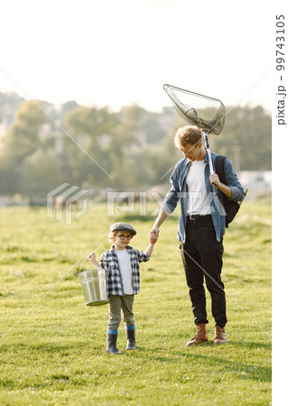 Dad and his child boy spend time outdoors together. Curly toddler boy wearing a plaid shirt and a hat. Boy holding a hand of his father. 99743105