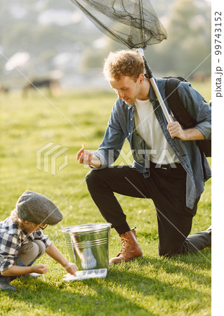 Dad and his child boy spend time outdoors together. Curly toddler boy wearing a plaid shirt and a hat. Boy standing near a bucket and listening his father. 99743152