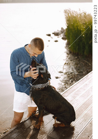 Handsome young male in casual outfit playing with cute dog while standing near the lake. Boy wearing blue shirt and white jeans shorts. Dog has a white and black scarf on his neck. 99743223