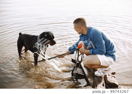 Handsome young male in casual outfit playing with cute dog while sitting near the lake. Boy wearing blue shirt and white jeans shorts. Dog has a white and black scarf on his neck. 99743267