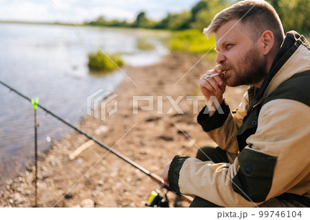 Close-up of pensive bearded fisherman wearing raincoat smoking sitting on travel chair at river bank waiting for catch, looking around. Concept of lifestyle, leisure activity on nature. 99746104